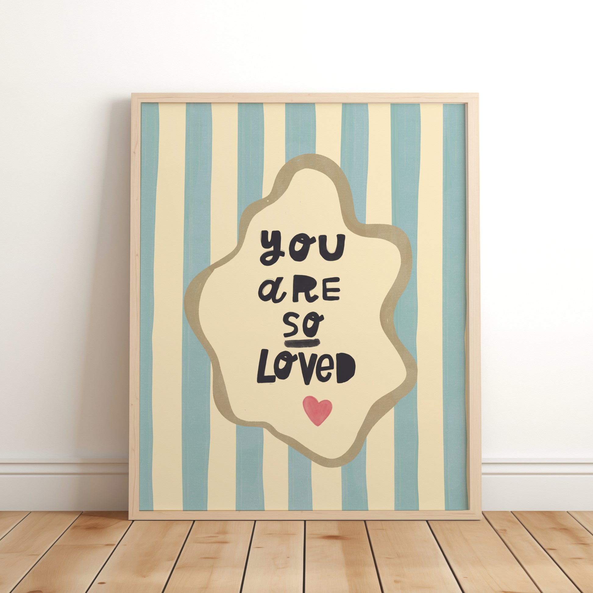 Nursery wall art print with the phrase “You Are So Loved” in playful black lettering, inside an organic cream frame with a gold outline, set over blue and cream vertical stripes and a small red heart.