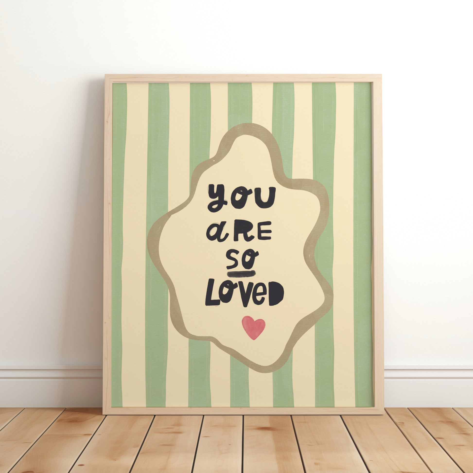Nursery wall art print with the phrase “You Are So Loved” in playful black lettering, inside an organic cream frame with a gold outline, set over green and cream vertical stripes and a small red heart.