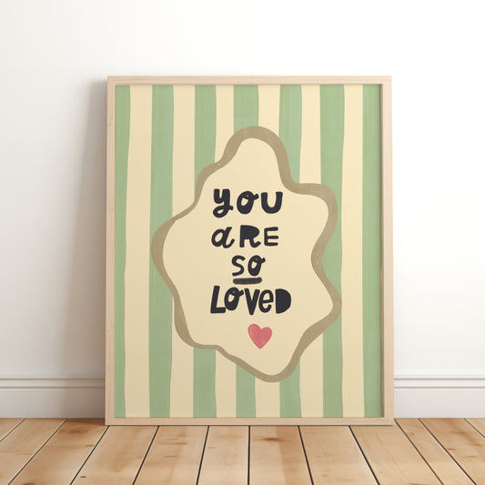 Nursery wall art print with the phrase “You Are So Loved” in playful black lettering, inside an organic cream frame with a gold outline, set over green and cream vertical stripes and a small red heart.
