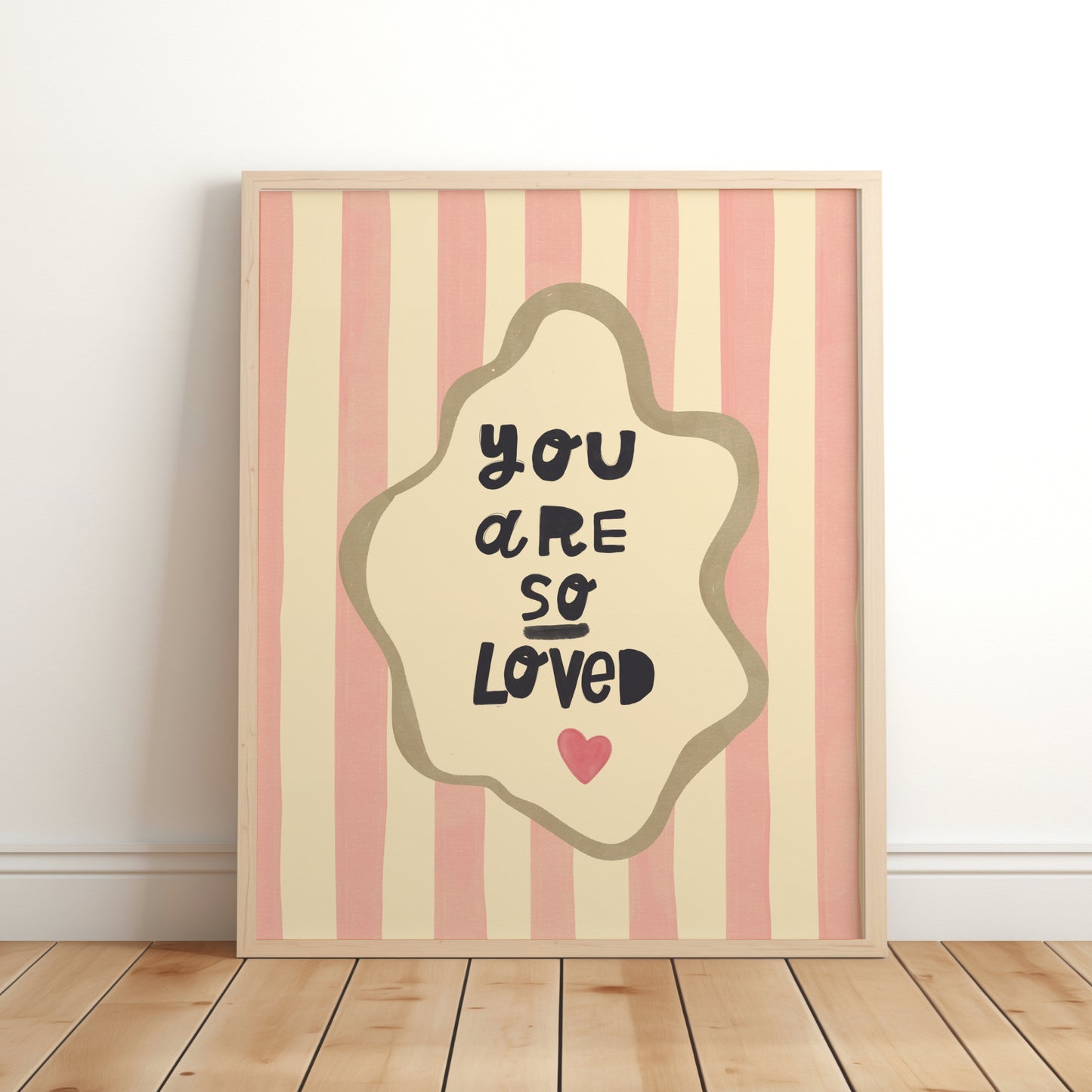 Nursery wall art print with the phrase “You Are So Loved” in playful black lettering, inside an organic cream frame with a gold outline, set over pink and cream vertical stripes and a small red heart.