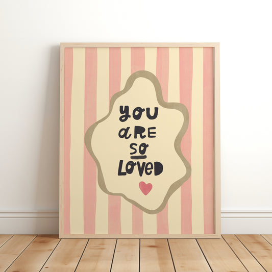 Nursery wall art print with the phrase “You Are So Loved” in playful black lettering, inside an organic cream frame with a gold outline, set over pink and cream vertical stripes and a small red heart.